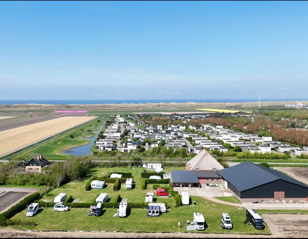 Luchtfoto van mini camping Hoeve Voorwaarts: caravans en campers op het groene terrein, met bloembollenvelden en de Noordzee op de achtergrond